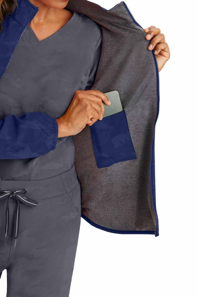 A young female Home Care Health Aide wearing a Purple Label Women's Destini Camo Scrub Jacket in Navy size Small featuring an interior cell phone pocket.