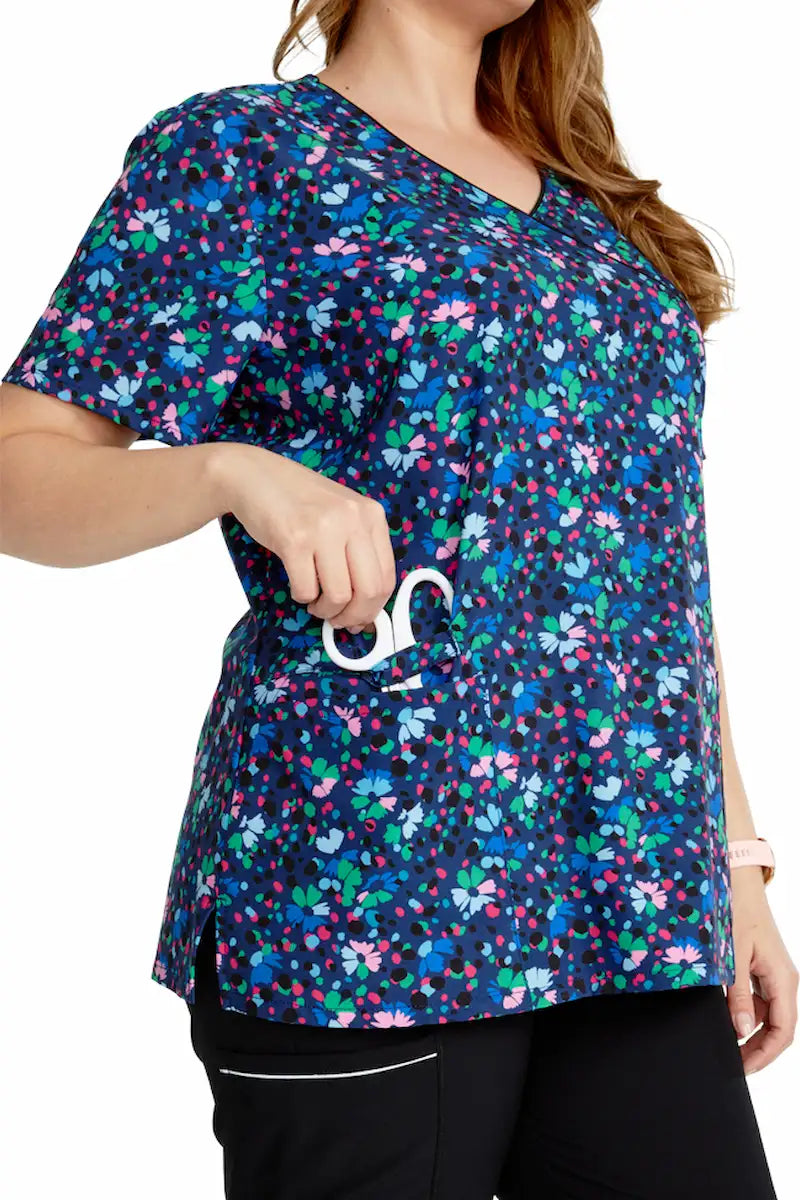 A young female Pediatric Nurse wearing a Cherokee Women's Mock Wrap Printed Scrub Top in "Confetti Daisies" featuring two spacious front patch pockets for additional on-the-job storage needs.