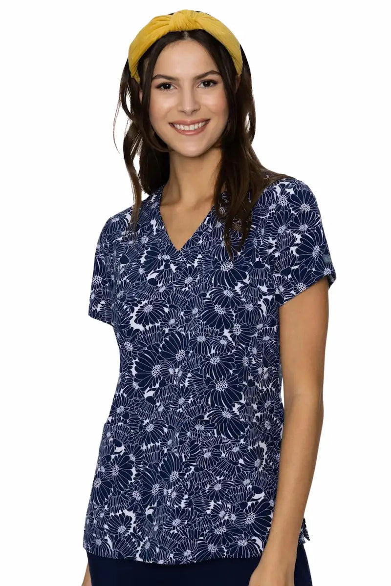 A young female Pediatric Nurse showcasing the front of the Med Couture Women's Vicky Print Scrub Top in Navy Daisy size medium featuring a classic V-neckline and two front patch pockets.