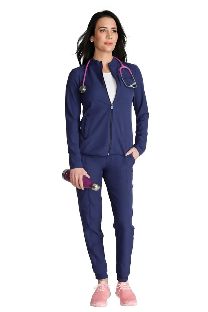 A full body shot of a young RN wearing a navy scrub uniform with an Allura Women's Zip-Up jacket over.