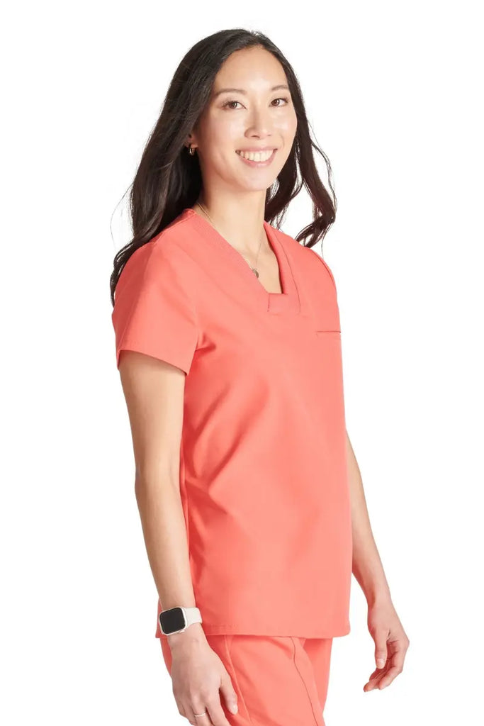 A young female Psychiatric Nurse wearing an Allura Women's Mitered V-Neck Scrub Top in Cayenne Pink size xs featuring a single welt chest pocket on the left side.