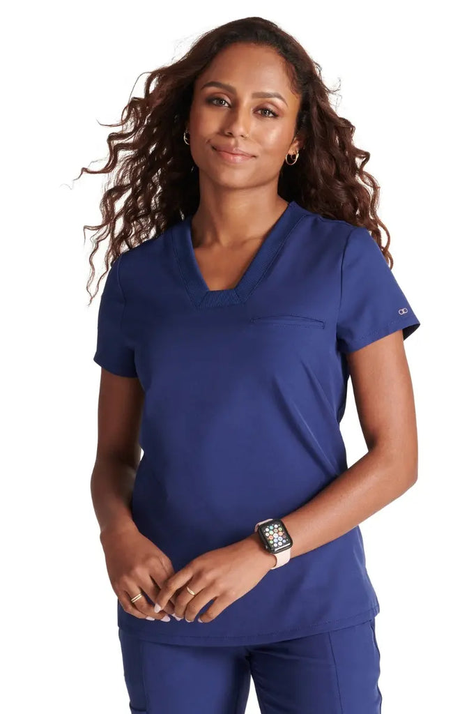 A young female LPN wearing an Allura Women's Mitered V-neck Scrub Top in Navy Blue size medium.