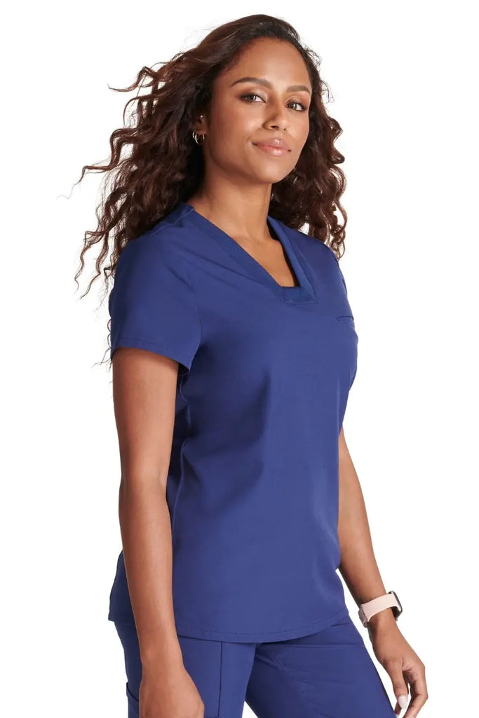 A young female Hospital Nurse wearing an Allura Women's Mitered V-Neck Scrub Top in Navy Blue size Medium featuring a single welt chest pocket on the left side.