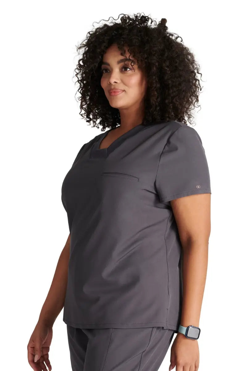 A female Nursing Assistant wearing an Allura Women's Mitered Scrub Top in Pewter featuring side vents.