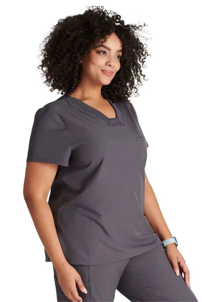 A young female Lab Tech wearing an Allura Women's Mitered V-Neck Scrub Top in Pewter size Medium featuring a single welt chest pocket on the left side.