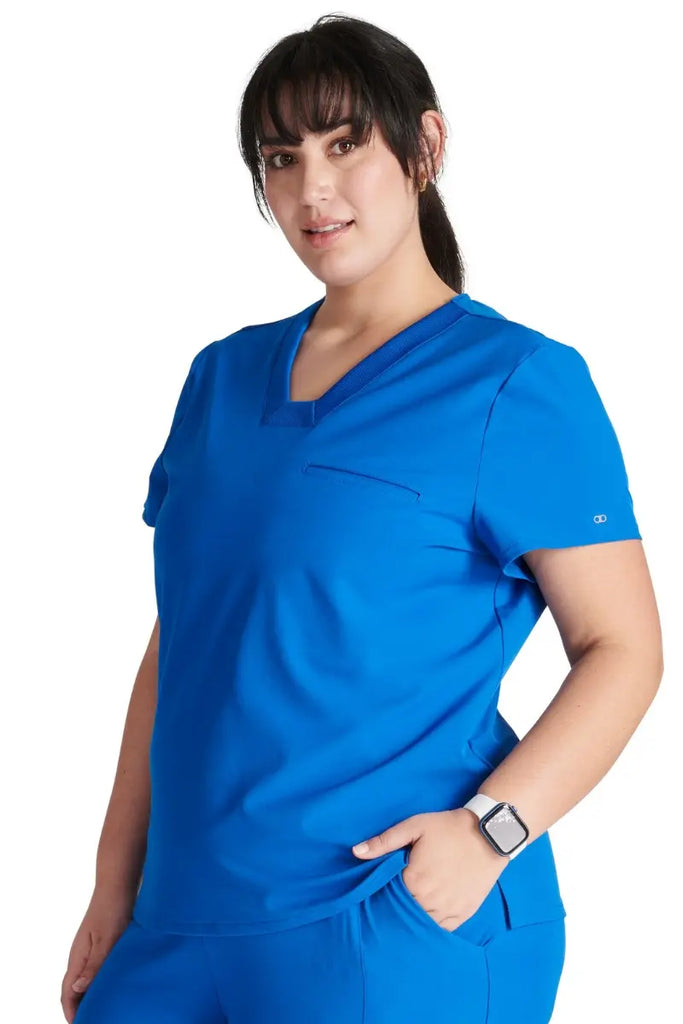 A young female Surgical Assistant wearing an Allura Women's Mitered V-Neck Scrub Top in Royal size Medium featuring a single welt chest pocket on the left side.