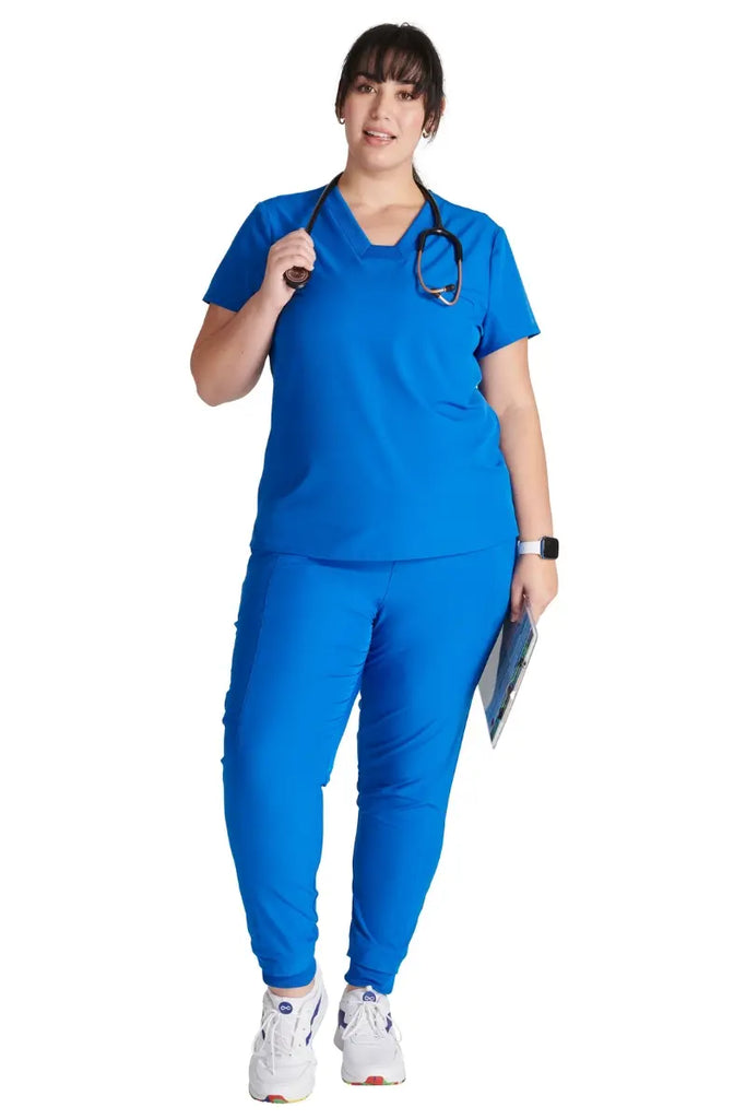 A full body shot of a young female Medical Assistant wearing an Allura Women's scrub uniform in Royal Blue size 3XL.