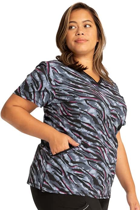 A young female RN wearing a Cherokee Infinity Women's Mock Wrap Print Top in "Wild for Tie Dye" featuring 2 front slash pockets.