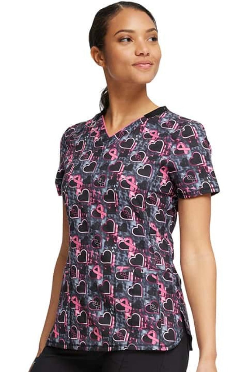 A female LPN wearing a Cherokee Infinity Women's Print V-neck Scrub Top in "Caring Beats" featuring front and back princess seams to ensure a flattering fit.