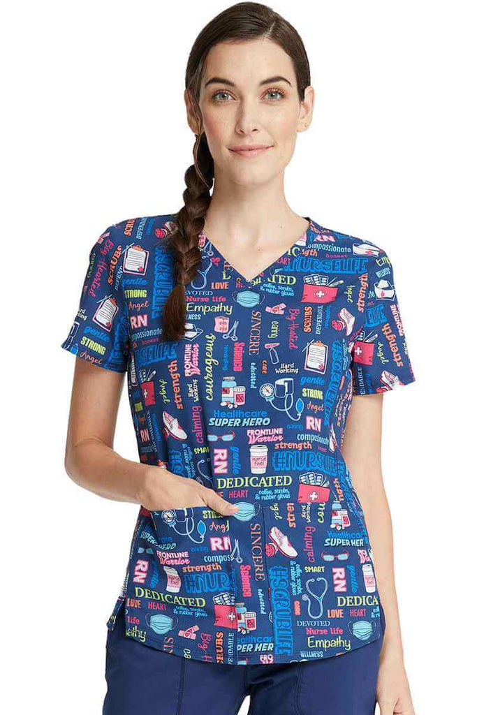 A young female RN wearing a Women's V-Neck Print Top from Cherokee Uniforms in "Scrub Life" featuring front vents. 