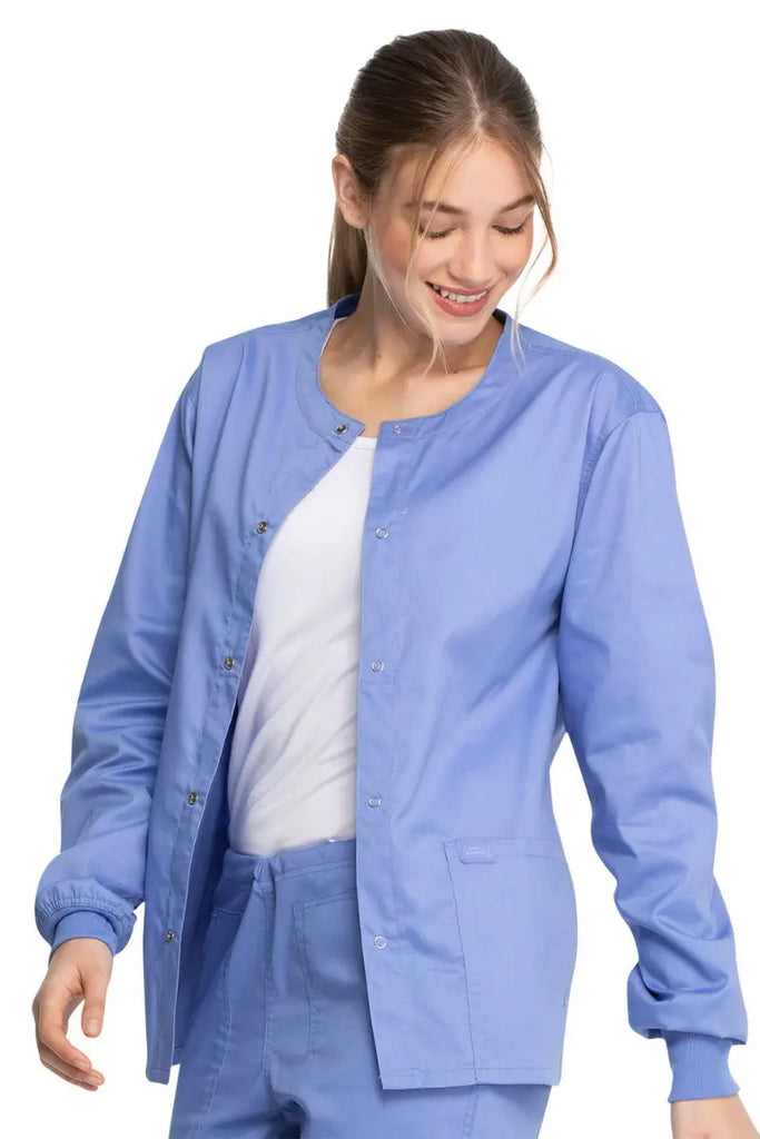 A young female Hospital Nurse wearing a Dickies Industrial Unisex Warm-Up Scrub Jacket in Ceil Blue size Large featuring a durable 80% polyester / 20% cotton stretch twill that can withstand daily wear and washing demands.