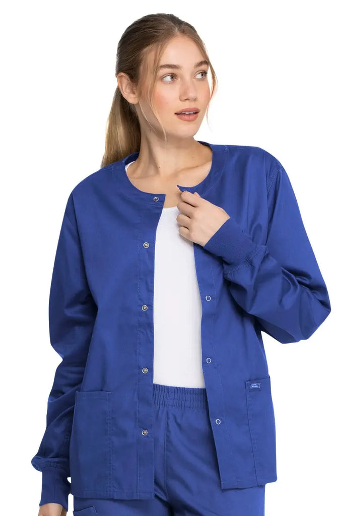 A young female CNA wearing a Dickies Industrial Unisex Warm-Up Scrub Jacket in Royal Blue size Large featuring a 5 metal button closure.