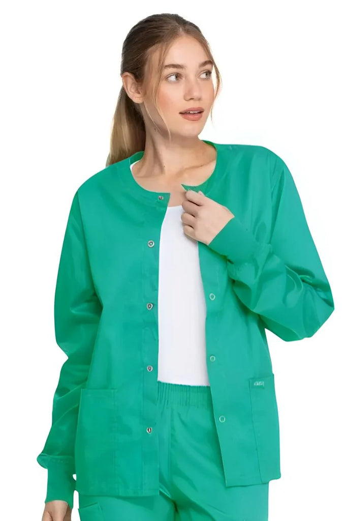 A young female Surgical Assistant wearing a Dickies Industrial Unisex Warm-Up Jacket in Surgical Green size XS featuring a 5 metal button closure.