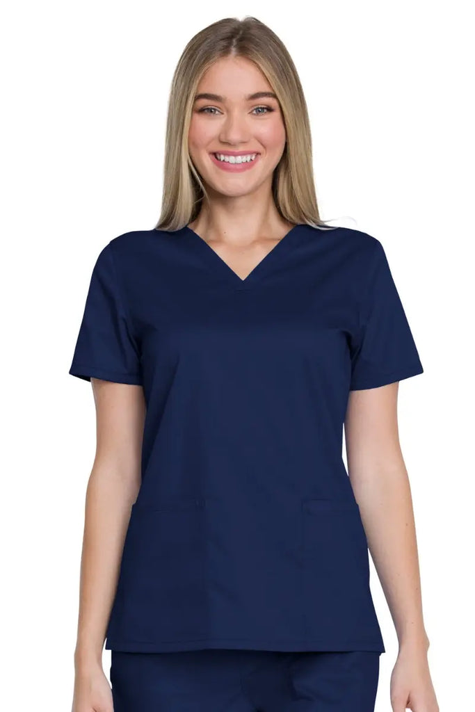 A young female Doctor wearing a Dickies Industrial Women's V-Neck Scrub Top in Navy Blue size XL featuring a durable 80% polyester/20% cotton stretch twill fabric that tackles industrial laundering easily.