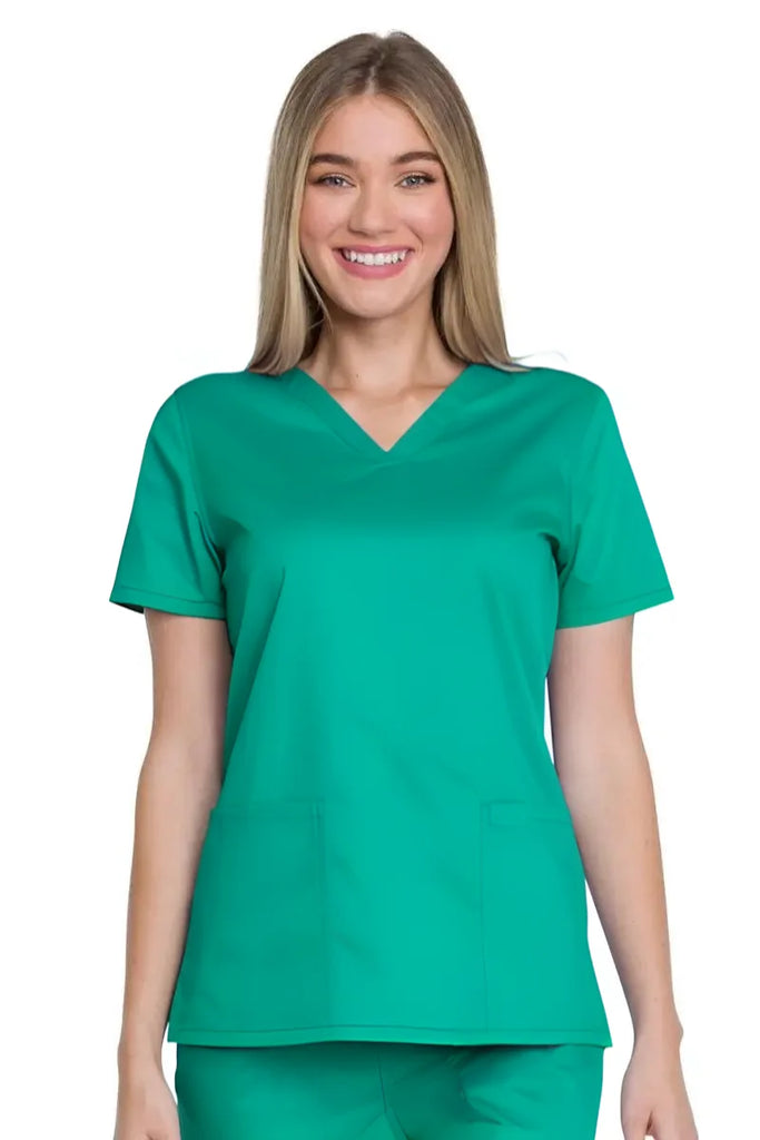 A young female Surgical Assistant wearing a Dickies Industrial Women's V-Neck Scrub Top in Surgical Green featuring a total of 3 pockets for ample storage space throughout the day.