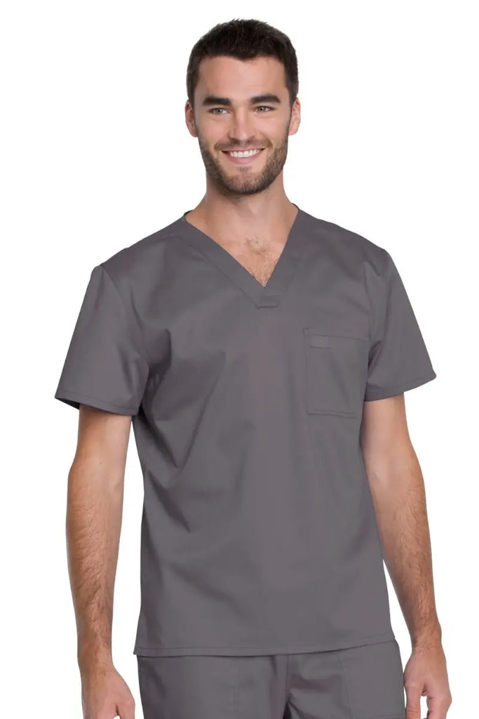 A young male Lab Technician wearing a Dickies Industrial Unisex Tuckable Scrub Top in Pewter featuring a V-neckline, short sleeves and one front chest pocket.