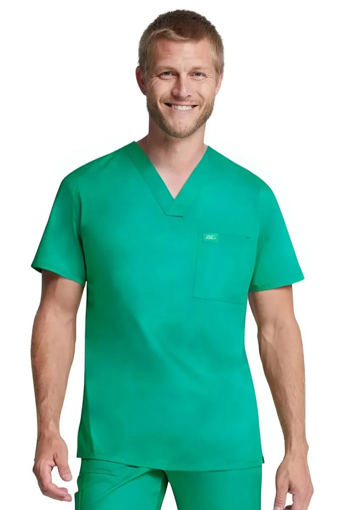 A young male Surgical Assistant wearing a Dickies Industrial Unisex Tuckable V-Neck Scrub Top in Surgical Green size 4XL featuring a conviently located chest pocket for on the go storage needs.