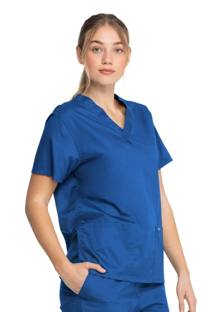A young female Certified Nursing Assistant wearing a Dickies Industrial Unisex V-Neck Scrub Top in Royal Blue size Medium featuring side slits for enhanced breathability and additional range of motion throughout the day.
