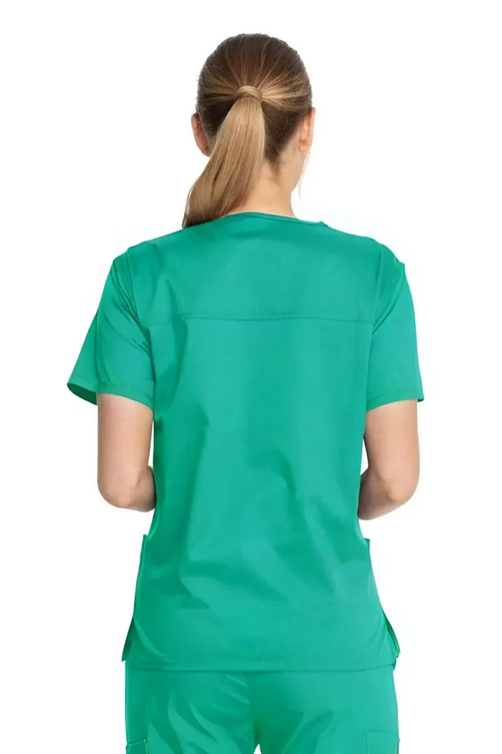 A young female First Assistant showcasing the back of the Dickies Industrial Unisex V-Neck Scrub Top in Surgical Green size Small featuring bartacks at stress points to provide extra strength and prevent tearing.