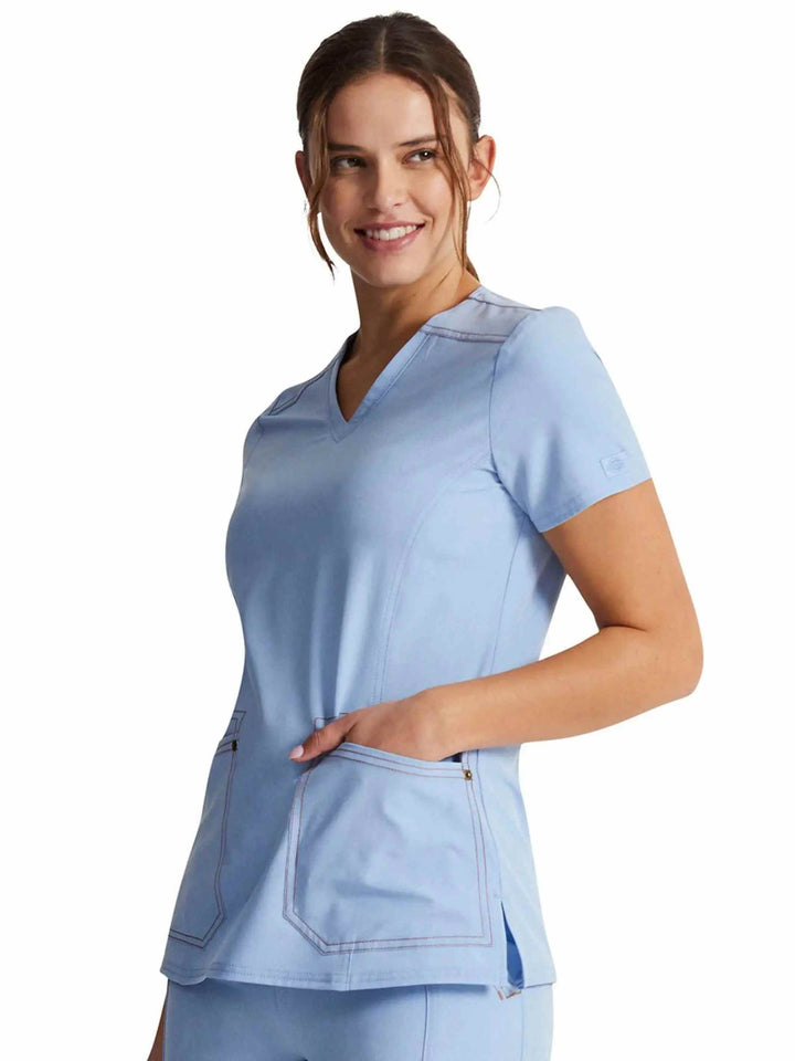 A young female Nurse wearing a Dickies Riveting Women's V-neck Scrub Top in Light Chambray featuring a curved hem and side slits.