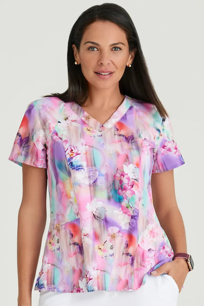 A young female Maternity Ward Nurse wearing a Barco One Women's Print V-Neck Scrub Top in Floral Blooms featuring two spacious front pockets, one inner right pocket, and a back left pocket.