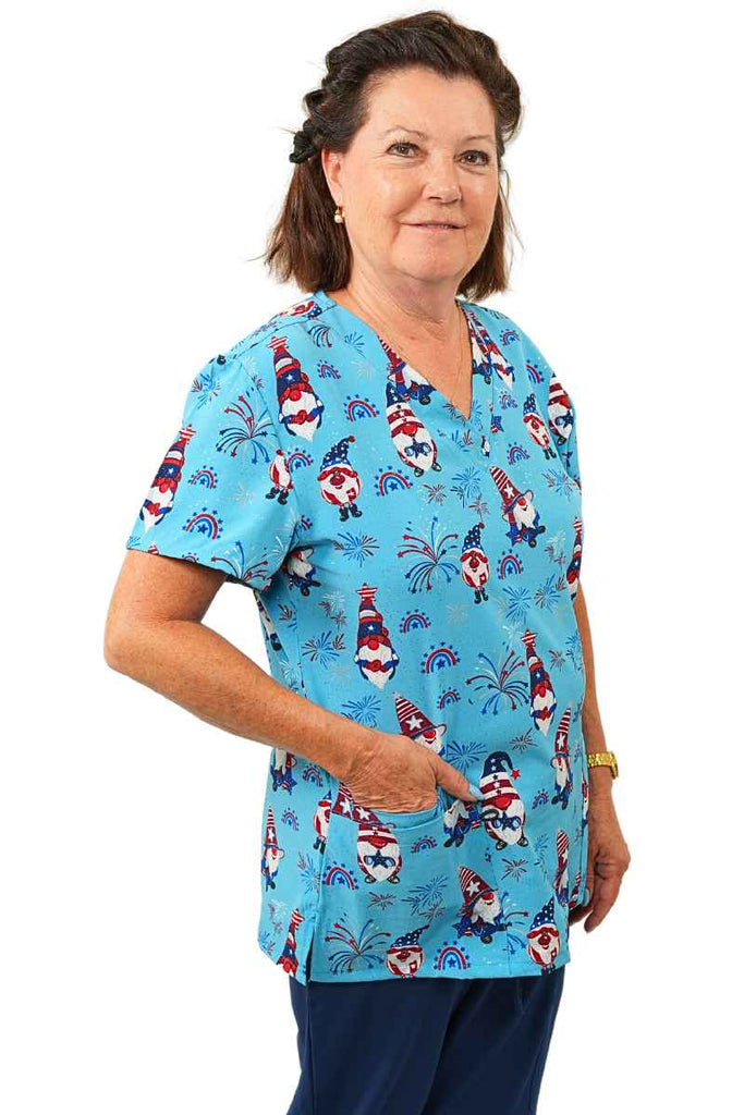 A young female Home Care Health Aide wearing a Luv Scrubs By MedWorks Women's Print Scrub Top in "Star Spangled Gnomes" size Small featuring short sleeves and a classic fit.