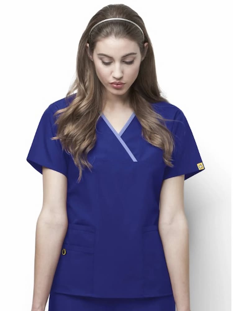 A young female PTA wearing a WonderWink Origins Women's Charlie Y-neck Scrub Top in Galaxy Blue size Medium featuring set-in sleeves & vented sides.