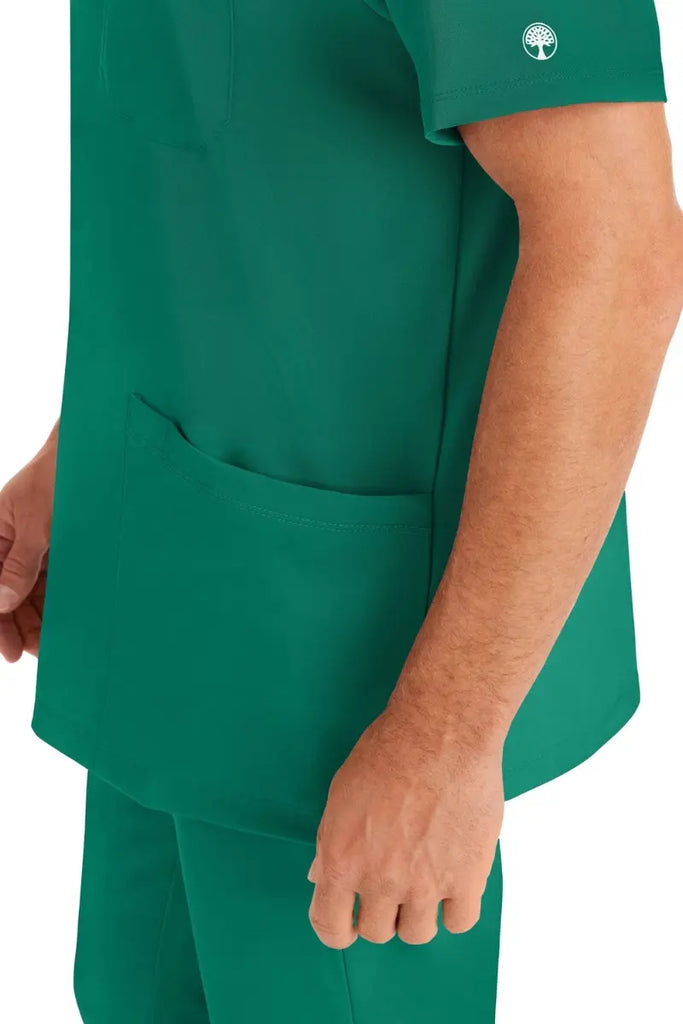 A young man wearing an HH-Works Men's Matthew V-Neck Scrub Top in Hunter Green featuring a quick drying, moisture wicking fabric.