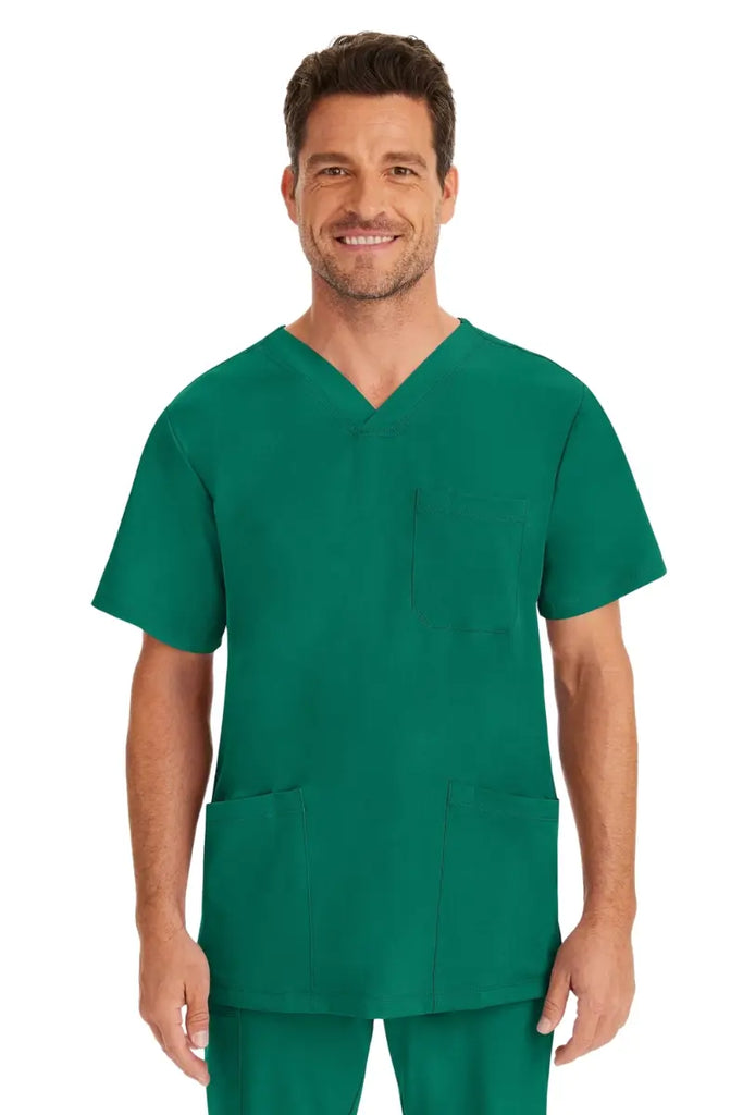 A young male Healthcare Hero wearing an HH-Works Men's Matthew V-Neck Scrub Top in Hunter Green featuring a v-neckline & short sleeves.