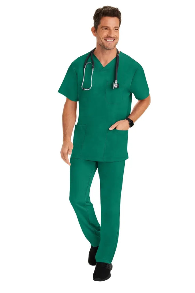 A young male Surgical Assistant wearing an HH-Works Men's Matthew V-Neck Scrub Top in Hunter Green featuring a fade resistant fabric to ensure a long-lasting comfortable fit.