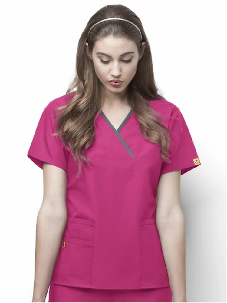 A young female pediatric nurse wearing a WonderWink Origins Women's Charlie Y-neck Scrub Top in Hot Pink size 2XL featuring a soft poly/cotton blend and enjoy ample storage space with the 5 total pockets.