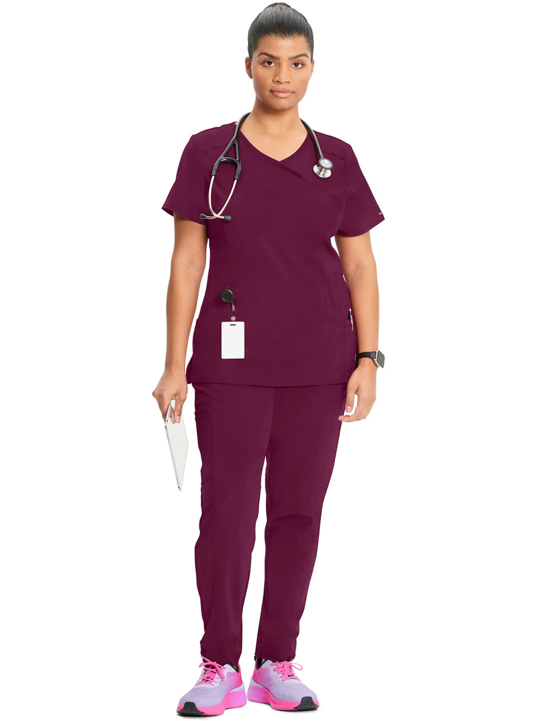 A full body shot of a nurse wearing the Infinity Women's Mock Wrap Scrub Top and Infinity Women's Mid-Rise Scrub Jogger in Wine on a white background.