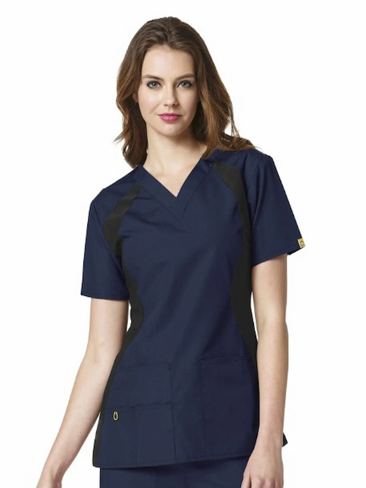 A young female Registered Nurse wearing a WonderWink Origins Women's Lima Knit Panel Scrub Top in Navy size XL featuring two front top-load pockets including one WonderWink signature triple pocket, signature ID bungee loop, hidden mesh pocket and utility loop.