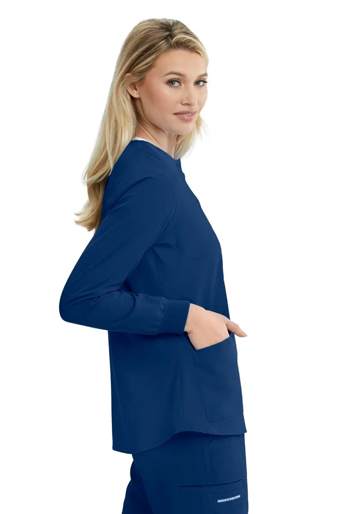 A young female Pharmacist showcasing the right side of the Skechers Women's Stability Snap Front Scrub Jacket in Navy size Medium featuring two front patch pockets.