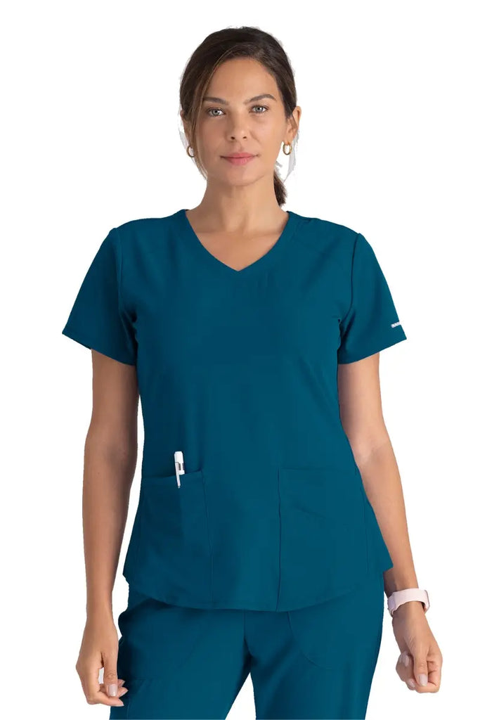 A young female Registered Nurse wearing a Skechers Women's Breeze V-neck Scrub Top in Bahama featuring two front patch pockets.
