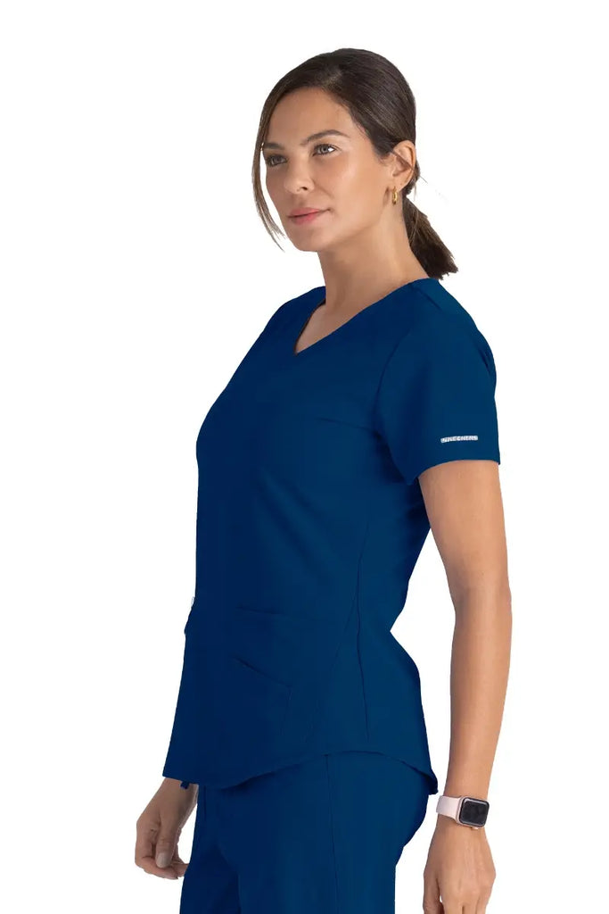 A young female LVN wearing a Skechers Women's Breeze V-neck Scrub Top in Navy size XS featuring the Skechers logo on the edge of the left side sleeve.