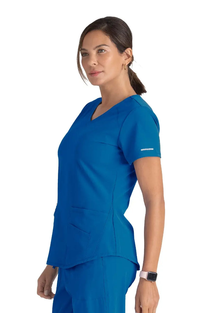 A young female LPN wearing a Skechers Women's Breeze V-neck Scrub Top in Royal size medium featuring the Skechers logo on the left sleeve.