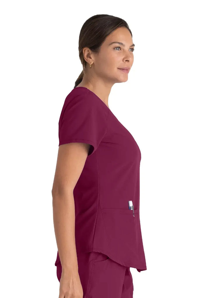A young female Nurse Practitioner wearing a Skechers Women's Breeze V-Neck Scrub Top in Wine size small featuring cover stitch detail throughout.