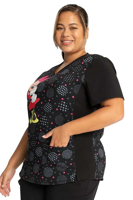 A young woman wearing a Cherokee Tooniforms Women's Stylized V-Neck Print Top in "Minnie Spotlight" featuring 2 front patch pockets for all of your on the go storage needs.