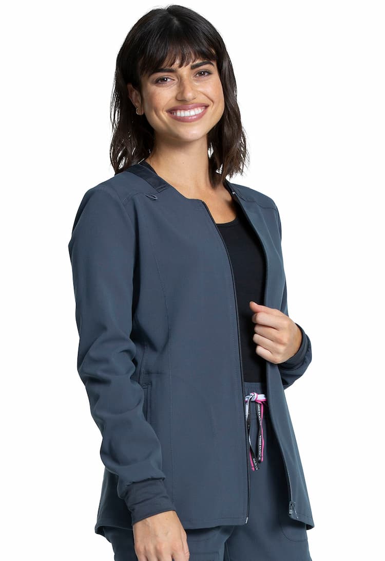 A young female Nurse Practitioner wearing a Vince Camuto Women's Zip Front Scrub Jacket in Pewter size Medium featuring a bungee ID badge loop on the wearer's right shoulder.