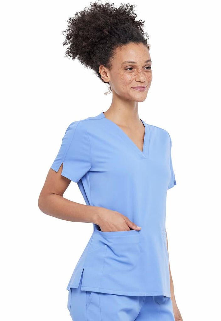 A young female Registered Nurse wearing a Vince Camuto Women's V-neck Scrub Top in Ceil Blue size XS featuring 2 front patch pockets.