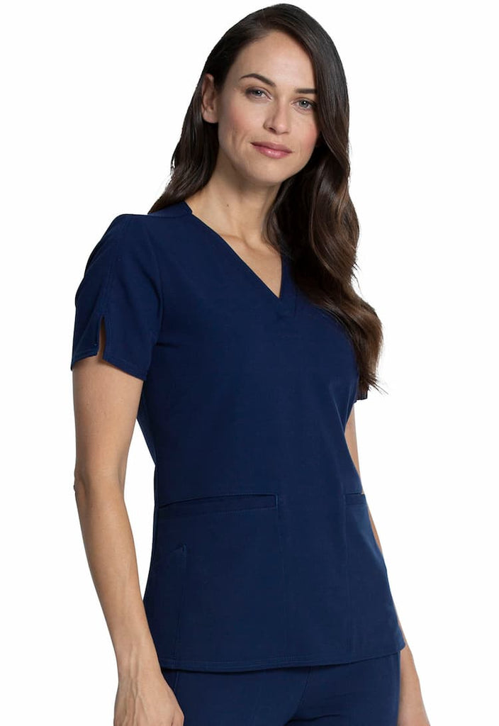 A young female Home Health Aide wearing a Vince Camuto Women's V-neck Scrub Top in Navy size 2XL featuring short sleeves with side slits for flair.