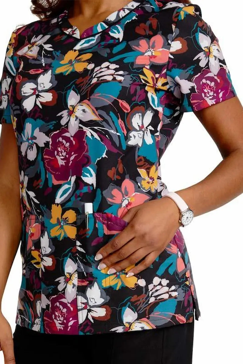 A young female pediatric nurse wearing a Cherokee Women's V-Neck Printed Scrub Top in "Artful Blooms" featuring two spacious front patch pockets for additional on the job storage space.