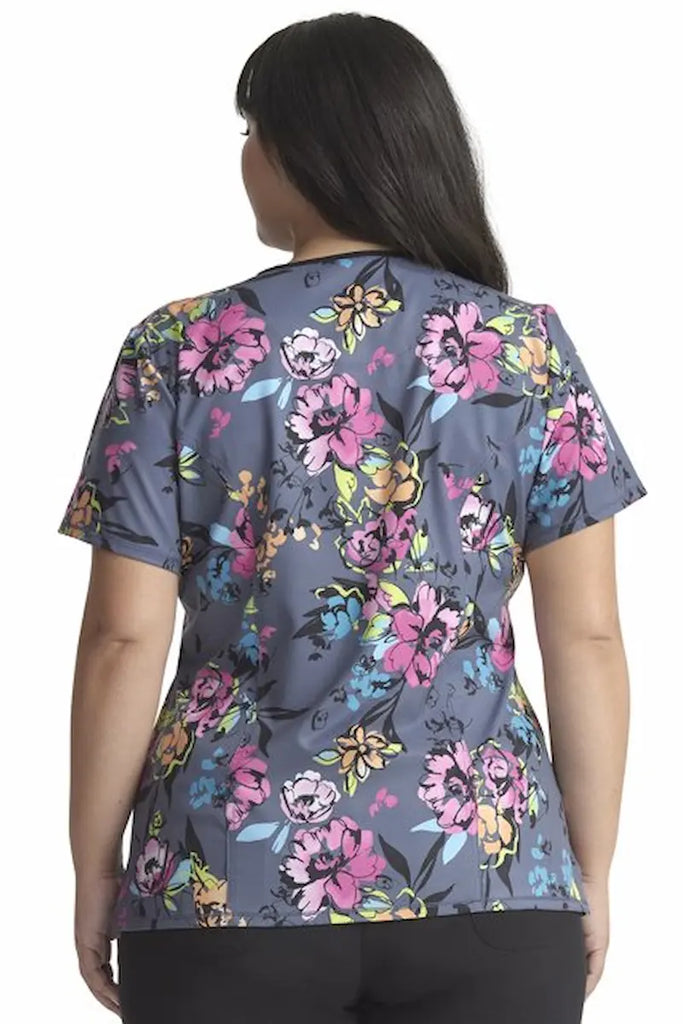 A young female Cherokee Infinity Women's Round Neck Printed Scrub Top in "Electric Blossoms" featuring a center back length of 26".