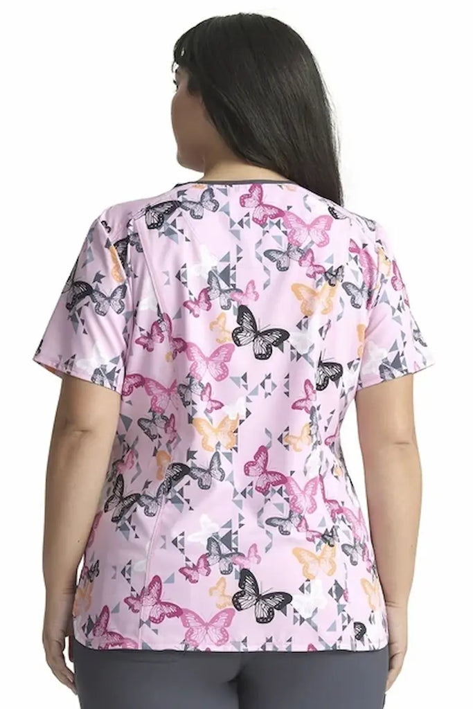 A young female Neonatal Nurse wearing a Cherokee Infinity Women's Print V-neck Printed Scrub Top in "Geo Flutter" size 3XL featuring an intricate geometric patterns and butterflies in a captivating color palette, adding a touch of personality and modern flair.