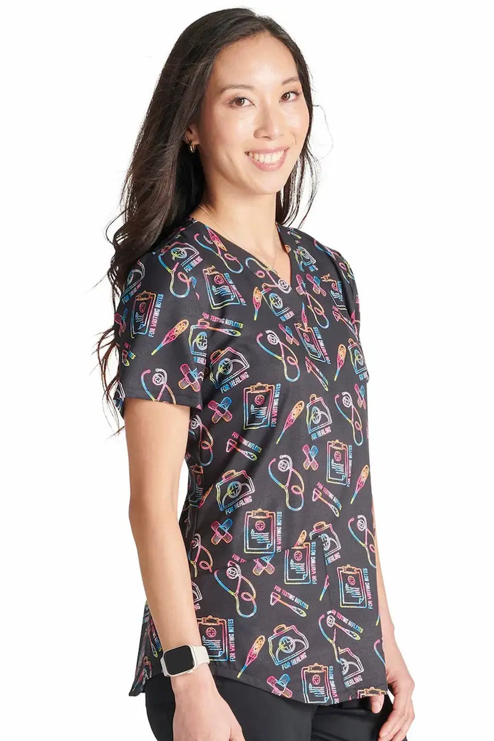 A young female children's nurse wearing a Cherokee Women's V-Neck Printed Scrub Top in Caring Essentials featuring short sleeves and a unique poly-cotton blended fabric to ensure a comfortable and breathable fit.