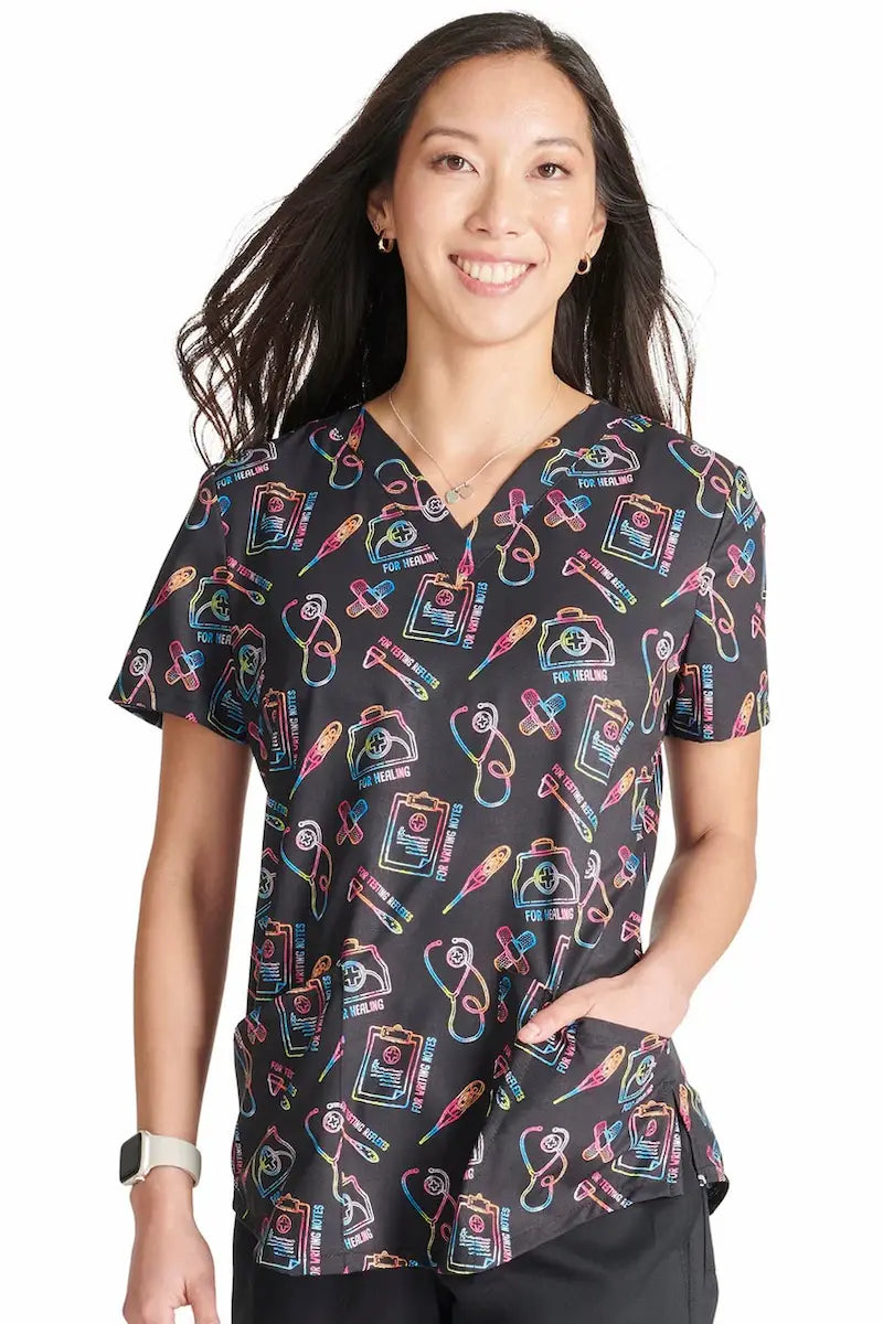 A young female Pediatric Nurse wearing a Cherokee Women's V-Neck Print Scrub Top in Caring Essentials featuring a rounded hem to provide a polished look all day long.