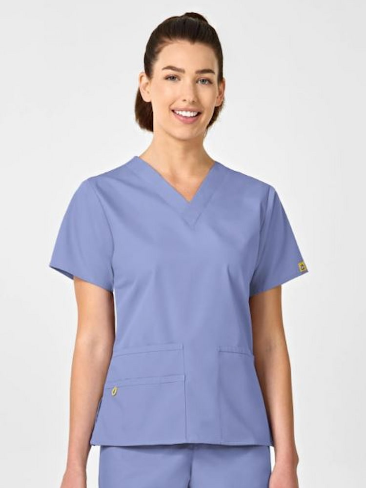 A young female Registered Nurse wearing a WonderWink Origins Women's Bravo V-neck Scrub Top in Ceil Blue size Medium featuring a hidden 3 part accessory loop.