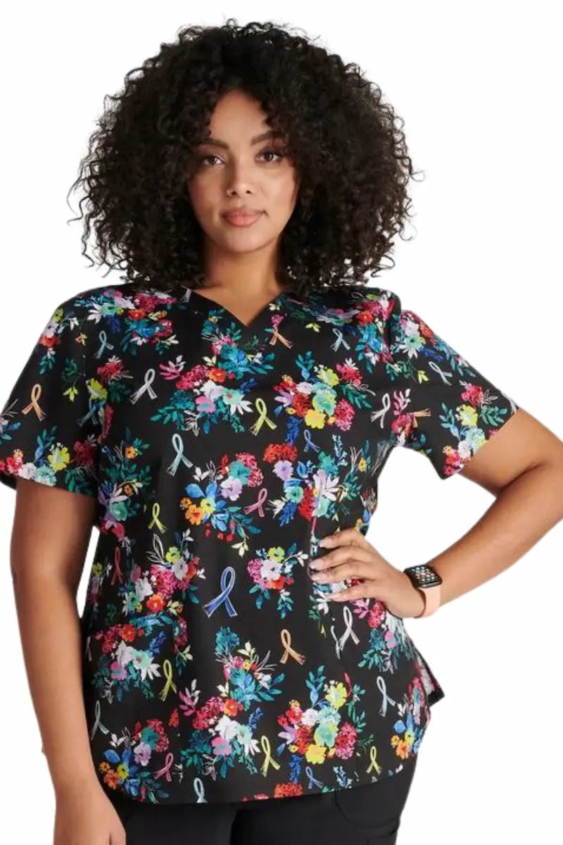 A young female nurse wearing the Hopeful Bouquets Cherokee Women's V-Neck Print Scrub Top in size 2XL on a solid white background.