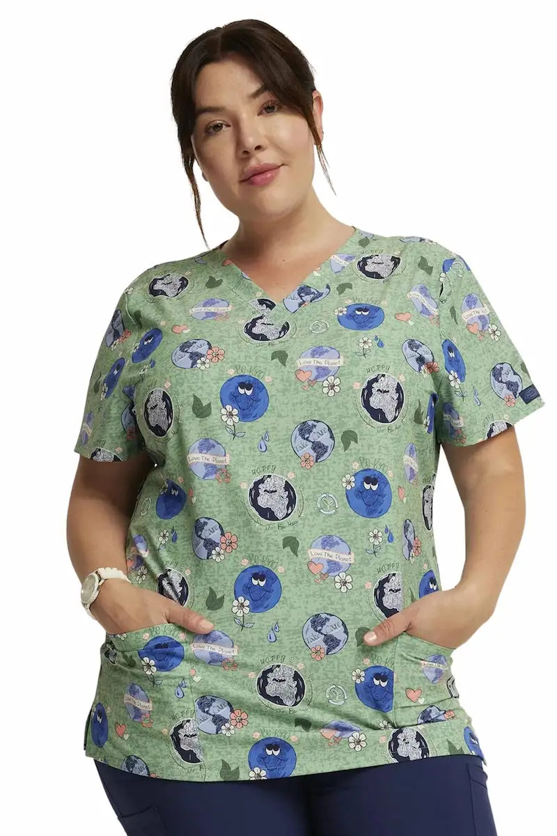 A young female Pediatric Nurse displaying the front of the Dickies Women's V-Neck Print Scrub Top in Happy to be Here size 2XL.